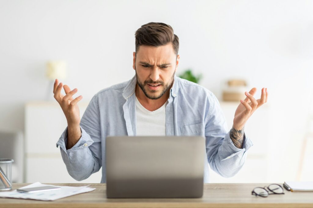 Angry millennial man looking at laptop screen and gesturing, having software, website error, sitting